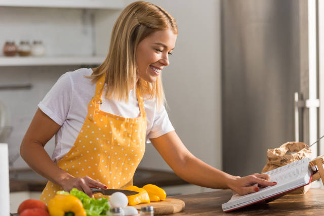 Smiling young woman in apron reading cookbook while cooking in kitchen